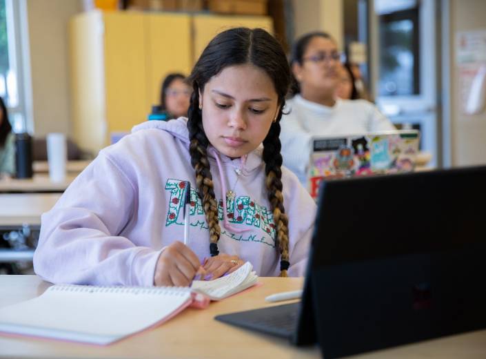 Early Childhood Development student writes in a notebook in her classroom. She is of Latinx descent and has thick dark braids and a nose ring.