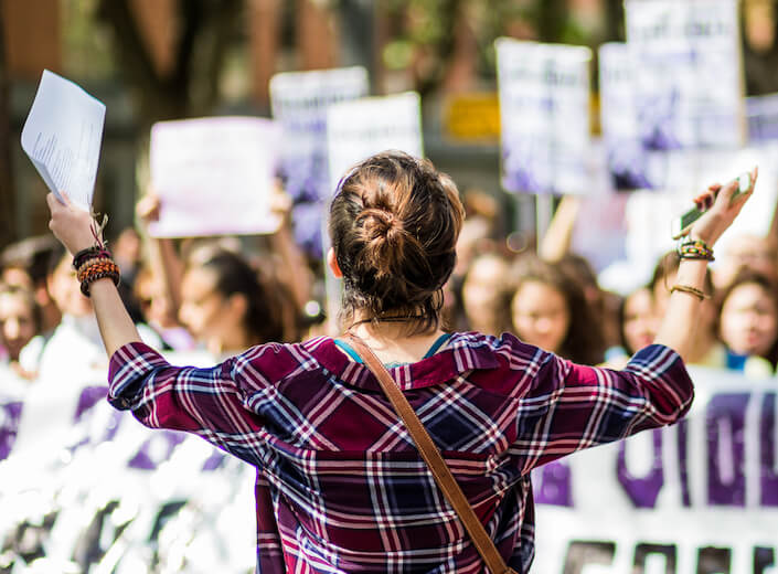 Young woman with light brown hair pulled up into a messy bun wears a red flannel and has both arms outstreched facing an out-of-focus crowd of protesters holdig signs.