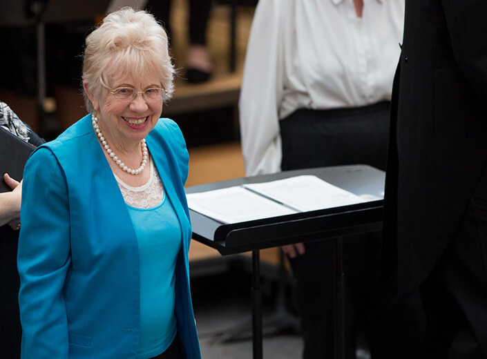 Woman in blue suit with light blonde hair, glasses, and a string of pearls smiles after a music performance.