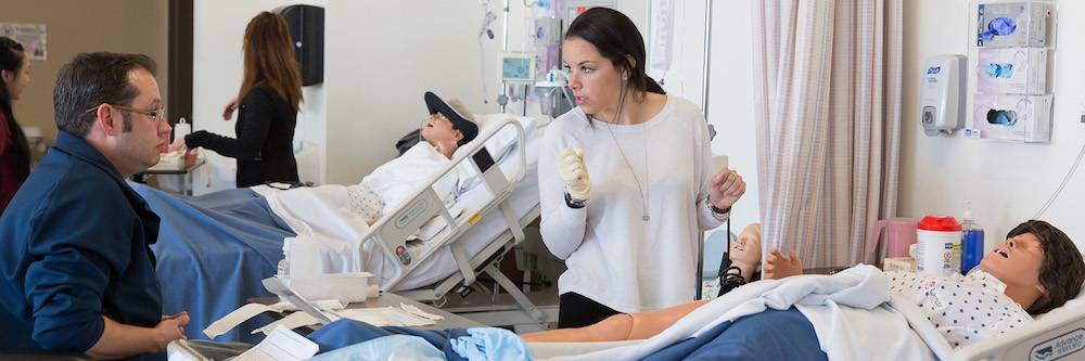 Nursing faculty works with student who is administering care in scrubs to a dummy patient in a hospital bed.