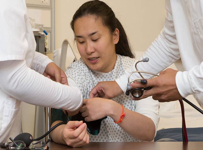 Practice patient, a young woman of Asian descent in a white hospital smock, allows student nurses to take her blood pressure in a hospital bed.