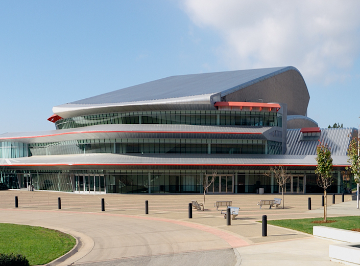 Phillips Hall on the SLO campus, a large modern-design building with with three stories. It has a metal sloping roof and red accents on each story. Large windows wrap around each level. It is photographed on a sunny day from outside.