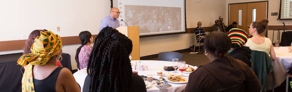 A professor leads a presentation on the Tuskeegee Study while attendees eat dinner in a large room on a college campus.