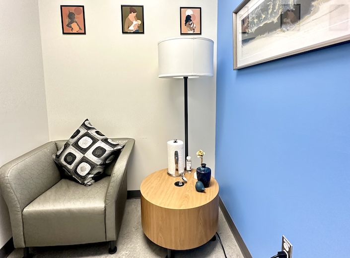 Lactation room with chair, table, lamp, and artwork featuring nursing mothers.