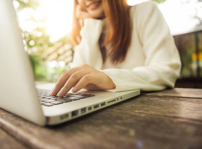 A young woman is pictured from the chin down. She has straight light brown hair, fair skin, and types with one hand on the keyboard of a laptop.