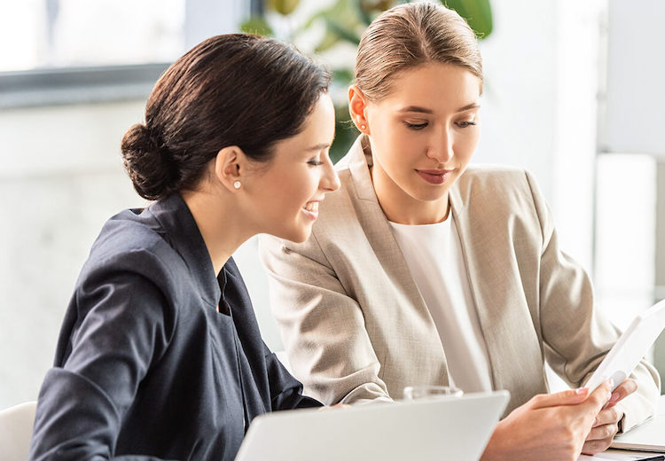 Two young women in office attire are working at a desk. One is blonde and the other has dark hair and tan skin.