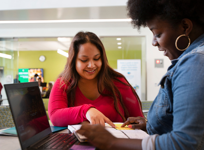 Two students are looking at a form. One has a laptop computer in her lap.