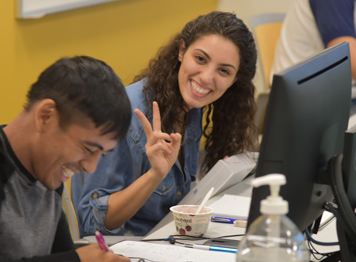 LatinX student is seating with her laptop at a table. She is looking up at another student who is also working at the table.