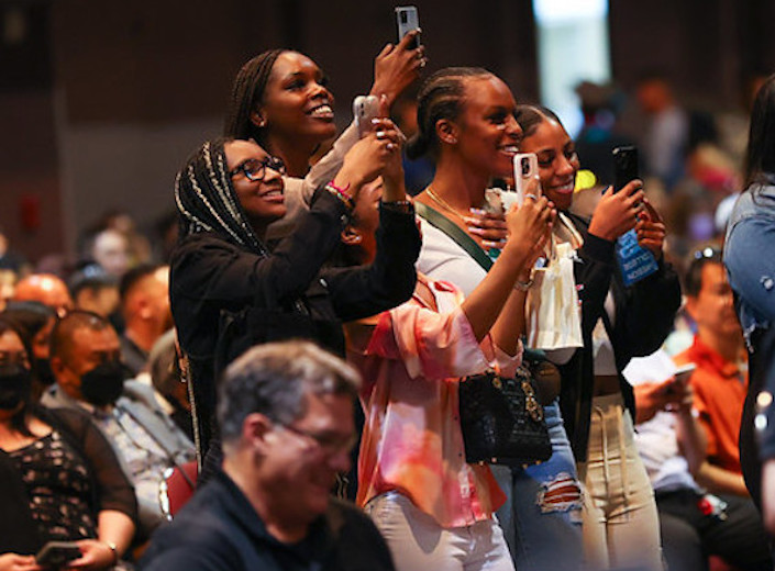 Women stand in audience of commencement ceremony. One wears a head scarf and glasses, two wear braided styles. They have brown skin.