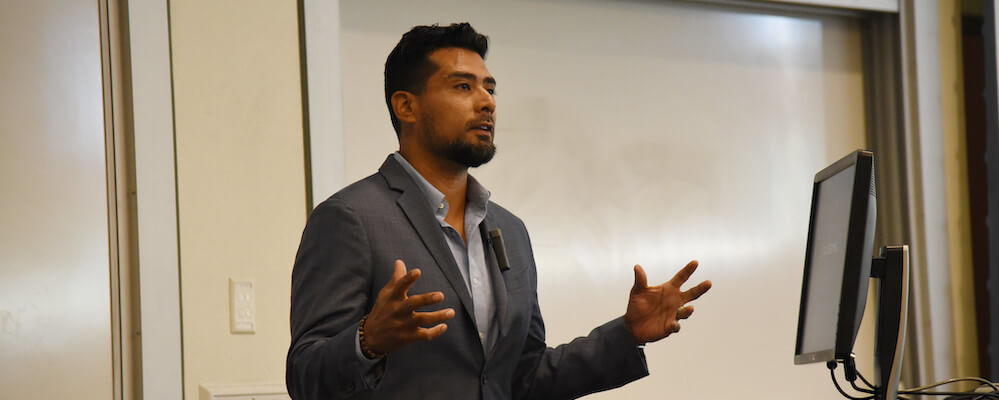 Dr. Victor Rios addresses a room of people in his grey suit. He is a sharp looking Latino man who looks youthful. He has short black hair and gestures with his hands.