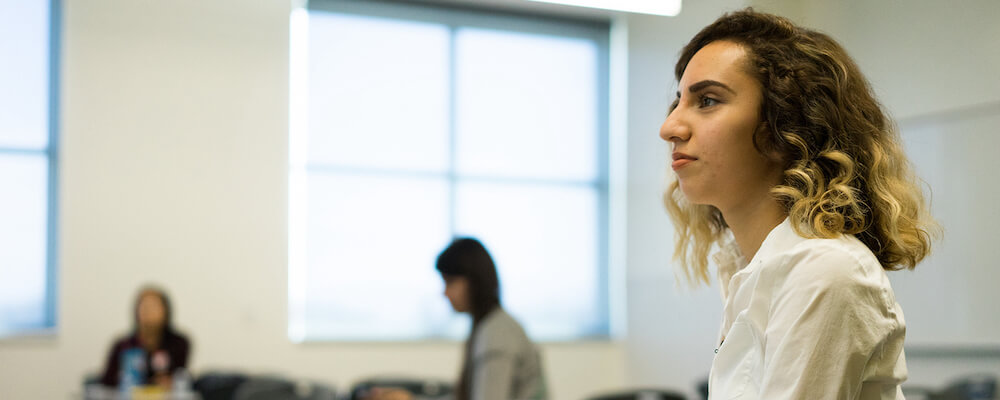 Young woman in classroom is pictured in profile. She has light brown curly hair to her shouders and olive skin. She wears a white button-down shirt.