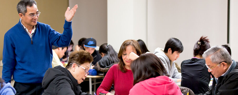 Male teacher in a blue sweater leads a class of adult ESL students on a college campus. The students work in small groups at round tables.