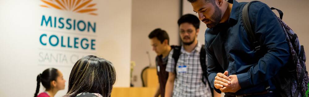 A young man leans over a table at a campus event to review information. He has dark hair and a blue sweate. In the background, a sign that reads "Mission College" is visible.