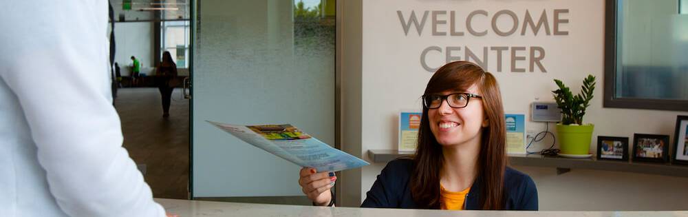 Welcome Center. A young woman with straight shoulder-length brown hair and glasses hands some papers to a student from behind a desk.