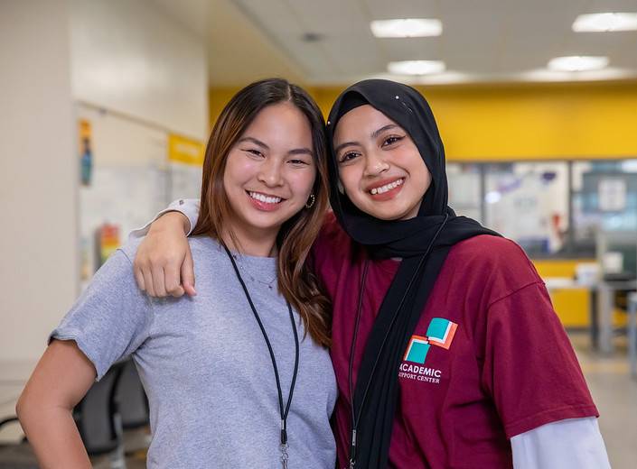 Two young women, one Asian-American, and the other Middle-Eastern in a head scarf smile and pose together.