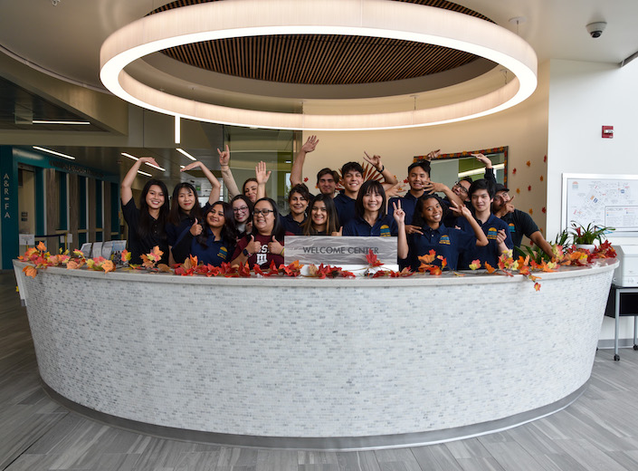 Welcome Ambassadors pose in a group wearing blue polo shirts behind the service desk in the Student Engagement Center. They are of many ethnicities and genders.