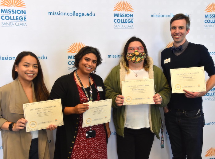 Vianey Topete holds an award/piece of paper with three colleagues in front of a backdrop decorated with the Mission College log.