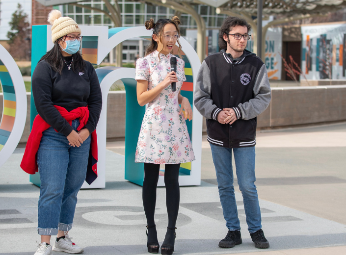 Casey Chang in light floral dress, black tights, addresses audience with microphone outside near Mission College sign on Central Plaza.